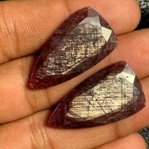 May include: Two faceted, elongated gemstones in a deep burgundy colour. The gems are triangular with a pointed end and a flat, polished surface. The interior of the stones has a shimmering, metallic appearance.