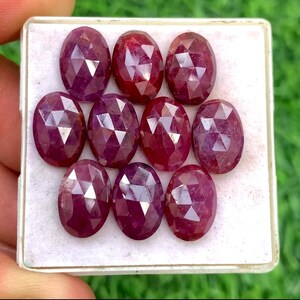Puede incluir: Una colección de nueve rubíes facetados de forma ovalada, cada uno con un tono rojo intenso. Las piedras preciosas están dispuestas en una bandeja rectangular blanca, mostrando sus superficies pulidas y cortes intrincados. Los rubíes están en una mano.