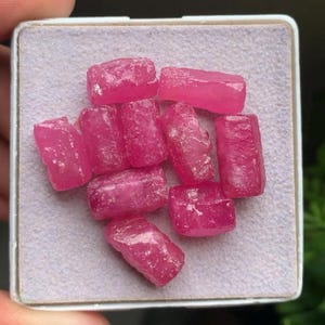 Pode incluir: Uma coleção de dez pequenas peças de pedras preciosas rosa vibrante, cortadas em bruto e dispostas em um grupo. Cada peça apresenta uma forma retangular irregular com uma superfície cristalina texturizada, exibida em uma almofada de espuma quadrada.