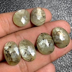 Puede incluir: Se muestran seis piedras preciosas facetadas, de forma ovalada y de color verde pálido. Las gemas tienen inclusiones internas y se sostienen en una mano. Las piedras tienen una superficie pulida y reflectante, y probablemente son para la fabricación de joyas.