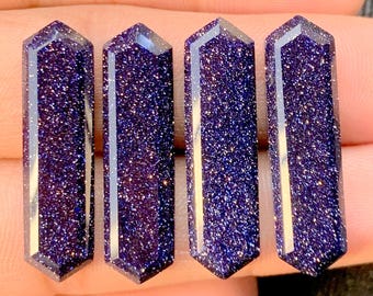 May include: Four blue sandstone crystal wands with a sparkling, glittery appearance. Each wand is a rectangular prism with a pointed top. The crystals have a deep blue color with flecks of gold throughout.