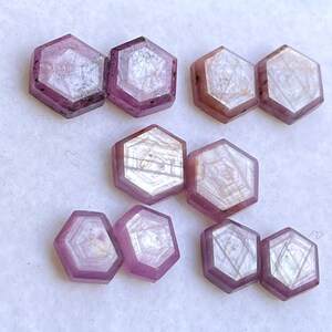 May include: A collection of hexagonal, polished gemstones in shades of purple and pink. The gems have a translucent quality, with internal patterns visible. The arrangement is on a white surface, showcasing the gems' facets and color variations.