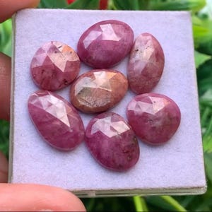 Könnte beinhalten: Eine Ansammlung von sechs ovalen, facettierten rosa Rubinen, die auf einem hellvioletten Präsentationskissen angeordnet sind. Jeder Edelstein besitzt eine polierte, mehrfach facettierte Oberfläche, die das Licht reflektiert.