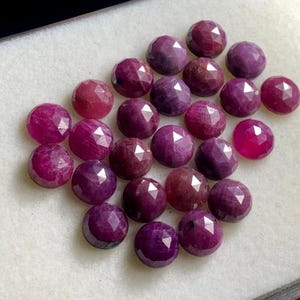 May include: A selection of faceted, round gemstones in deep red and purple hues. The gems are displayed on a white surface, highlighting their polished surfaces and geometric cuts. The gemstones vary in colour intensity.