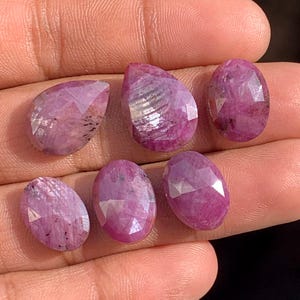 May include: Six faceted, oval and pear-shaped ruby gemstones in varying sizes. The rubies display a deep pinkish-red hue with visible internal inclusions. The gemstones are displayed on a light-toned background.