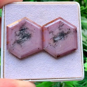 Könnte beinhalten: Zwei sechseckig geschliffene rosa Edelsteine mit sichtbaren dunkelgrauen und schwarzen mineralischen Einschlüssen auf der Oberfläche. Die Steine haben eine flache, polierte Oberseite und liegen nebeneinander auf einem hellen, strukturierten Quadrat.