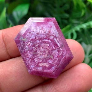 May include: A hexagonal, faceted gemstone with a vibrant magenta and pink hue. The surface displays a complex, layered geometric pattern with visible internal inclusions and a semi-translucent quality, showcasing a distinct six-sided crystal structure.