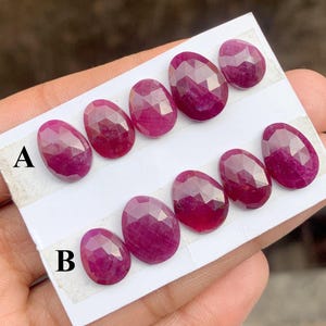 Puede incluir: Colección de gemas de rubí facetadas de forma ovalada. Los rubíes son de un color rojo intenso y rico, dispuestos sobre una tarjeta blanca. La tarjeta tiene las letras "A" y "B" impresas.