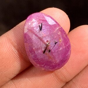 Peut inclure: Une pierre précieuse de rubis ovale et facettée. La gemme est d'une couleur rose-rouge vif avec des inclusions sombres. La pierre est tenue dans une main, mettant en valeur sa surface polie et sa beauté naturelle. Idéal pour la joaillerie.