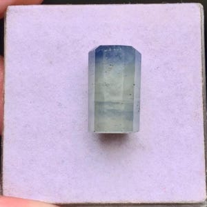 May include: A faceted gemstone with a cylindrical shape, displaying a colour gradient that transitions from a deep blue at the top to a pale, translucent off-white or light grey at the base, featuring visible internal inclusions and facets.