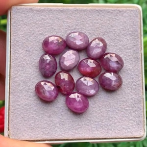 May include: A collection of eleven oval-shaped, faceted rubies with a deep pinkish-red hue. Each gemstone features a polished surface with geometric facets that reflect light, arranged in a cluster on a light grey, textured display pad.