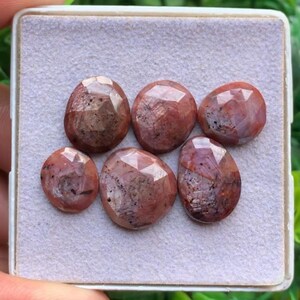 Puede incluir: Cinco piedras preciosas facetadas de forma ovalada con un tono marrón rojizo e inclusiones oscuras. Las piedras están dispuestas en un grupo sobre una almohadilla texturizada de color gris claro dentro de un recipiente cuadrado blanco.