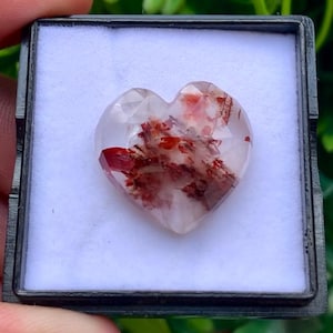 May include: A heart-shaped faceted gemstone with a translucent, clear base featuring internal red and dark brown mineral inclusions. The stone is displayed inside a square black plastic case with a white velvet-like insert.