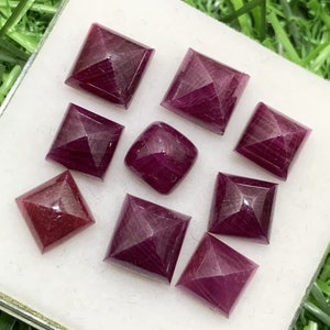 May include: Nine loose, faceted, square-shaped, ruby red gemstones. The gemstones are arranged in a random pattern on a white surface.