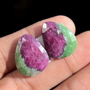 Może przedstawiać: Dwa fasetowane kamienie szlachetne w kształcie łzy. Kamienie szlachetne charakteryzują się żywą mieszanką kolorów, z głębokim, purpurowym środkiem i zieloną obwódką. Szlif kamieni tworzy wiele odbijających światło powierzchni.