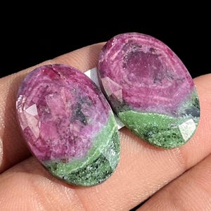Könnte beinhalten: Zwei ovale Edelsteine mit facettierter Oberfläche. Die Steine weisen eine lebendige Farbkombination auf, darunter tiefes Rosa, Grün und Schwarz. Die Edelsteine werden in der Hand einer Person gehalten.