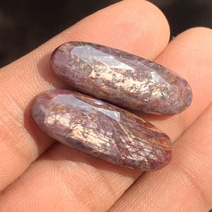 Pode incluir: Duas pedras preciosas alongadas e facetadas com um tom roxo profundo e brilho iridescente. As pedras são seguradas em uma mão, mostrando suas superfícies polidas e variações naturais. As gemas têm uma forma única e orgânica.