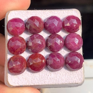 May include: A collection of twelve faceted, round-cut rubies presented in a white tray. The gemstones are a deep red hue, with visible internal patterns. Each ruby is multi-faceted, reflecting light. The gems are arranged in three rows of four.