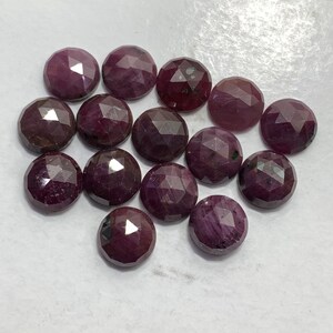 Puede incluir: Una colección de 14 piedras preciosas de rubí facetadas, cada una con un tono rojo intenso y una forma ligeramente irregular. Las piedras están dispuestas en un grupo suelto sobre una superficie blanca.