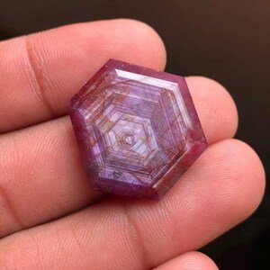 May include: A hexagonal-shaped ruby gemstone featuring a distinct trapiche pattern with radiating lines and concentric geometric bands in shades of deep pink and red, displaying a polished surface with visible internal inclusions and natural textures.
