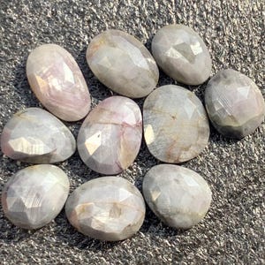 Op de afbeelding: Een verzameling van negen gefacetteerde, ovaalvormige edelstenen. De stenen hebben een lichtlavendelkleur met interne adering en een gepolijst, reflecterend oppervlak. De edelstenen liggen op een getextureerd, donkergrijs oppervlak.