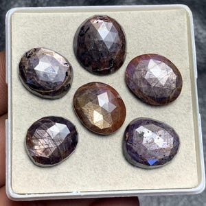 Può includere: Sei gemme ovali sfaccettate in un vassoio bianco. Le gemme presentano una gamma di colori, tra cui viola scuro, marrone e oro, con superfici riflettenti. Le pietre sono disposte in un motivo rettangolare.