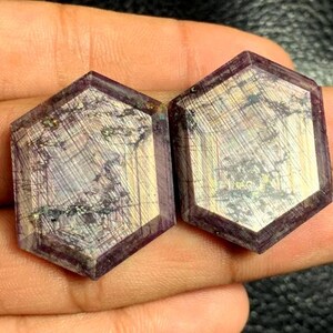 Peut inclure: Deux pierres précieuses hexagonales d'un violet profond, avec des motifs internes complexes. Les pierres ont une surface polie avec des couleurs or et grise, créant une texture visuelle unique. Les pierres sont tenues dans une main.