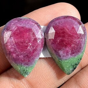 Può includere: Due pietre preziose di rubino sfaccettate a forma di pera. Le gemme hanno un centro rosso intenso, che sfuma in una tonalità viola e termina con una base verde. Le superfici sfaccettate riflettono la luce.