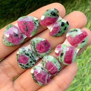 Peut inclure: Plusieurs pierres précieuses de rubis zoïsite facettées. Les pierres présentent un mélange de couleurs vives, avec des teintes roses et rouges profondes contrastant avec des mouchetures vertes et noires. Les pierres sont tenues dans une main sur un fond vert flou.