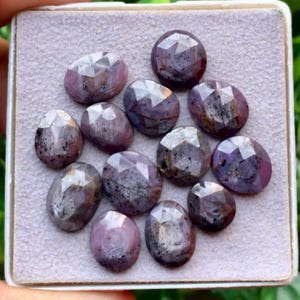May include: A collection of twelve faceted, oval-shaped gemstones in shades of dusty purple and grey. Each stone features a rose-cut top with multiple triangular facets, displaying natural inclusions and dark speckles throughout the surface.