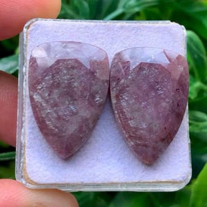 May include: Two pear-shaped, faceted gemstones with a deep mauve and dusty rose color palette. The stones feature a translucent quality with visible internal inclusions and a polished surface, displayed side-by-side on a light purple foam insert.