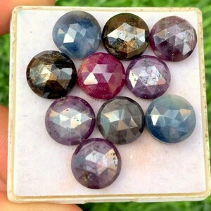 Peut inclure: Assortiment de neuf pierres précieuses facettées dans une variété de couleurs, notamment le bleu, le violet, le rouge et le noir. Les pierres précieuses sont présentées dans une boîte blanche.