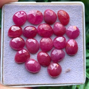 Può includere: Una collezione di diciassette rubini sfaccettati di forma ovale con una profonda tonalità rosso rosato. Ogni gemma presenta una superficie lucida con molteplici sfaccettature triangolari disposte in un motivo geometrico, su un vassoio in feltro grigio chiaro.