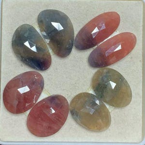 Könnte beinhalten: Acht ovale Edelsteine mit facettiertem Schliff. Die Edelsteine sind in den Farben Pink, Rot, Braun und Grün gehalten.