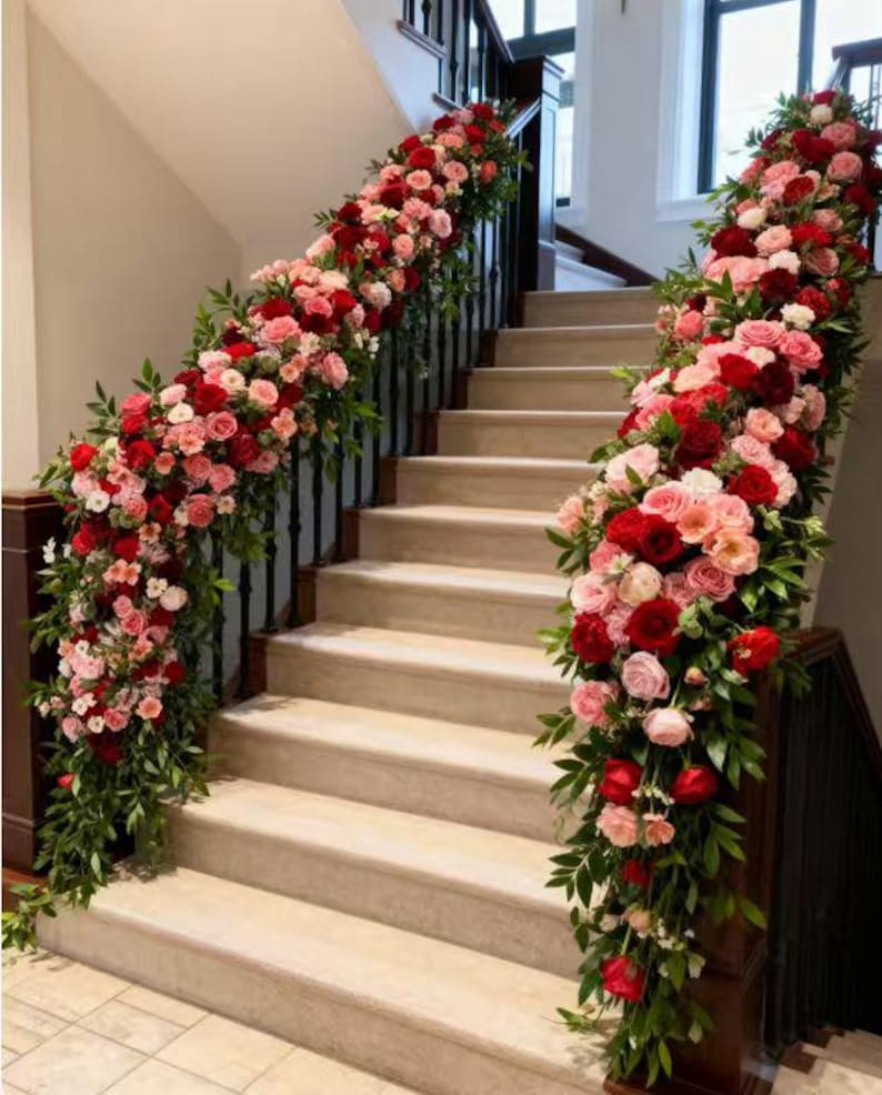 May include: A staircase adorned with vibrant floral arrangements. Lush garlands of red and pink roses, interspersed with greenery, cascade along the banisters. The steps are a neutral beige, creating a striking contrast with the colorful flowers.