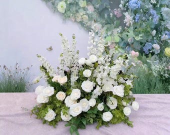 以下が含まれることがあります： 白いバラやその他の白い花をあしらった豪華なフラワーアレンジメント。緑の葉がアクセントになっています。パステルカラーの花と緑を背景に、柔らかくロマンチックな雰囲気を演出しています。