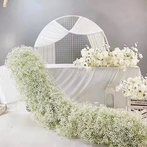 May include: A white wedding ceremony backdrop featuring a white metal grid archway draped with white fabric. A long garland of white baby's breath flowers is draped across the front of the table. The table is covered with a white tablecloth. Two white wooden boxes with the text "Wooden Box No.2" are on the table.
