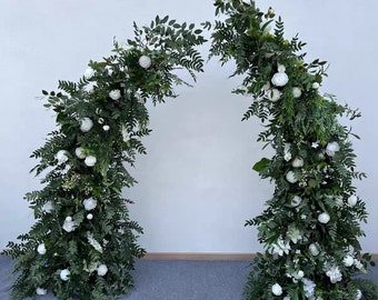May include: A wedding arch made of greenery and white flowers. The arch is made of two separate pieces, each with a curved shape. The arch is decorated with white flowers and green leaves.