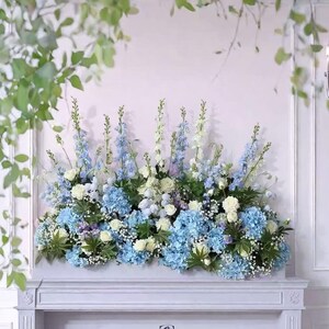 Puede incluir: Arreglo floral con hortensias azules, rosas blancas y flores azules y blancas esbeltas. El arreglo está colocado sobre una repisa blanca, con follaje en el fondo. La estética general es elegante y fresca.