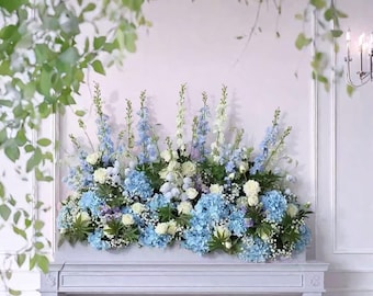 Puede incluir: Arreglo floral con hortensias azules, rosas blancas y flores azules y blancas esbeltas. El arreglo está colocado sobre una repisa blanca, con follaje en el fondo. La estética general es elegante y fresca.