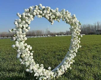 May include: A white floral heart-shaped archway made of artificial flowers. The archway is standing in a grassy field.