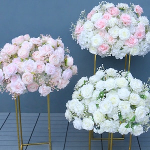 Puede incluir: Tres soportes de metal dorado con tapas redondas, cada uno con un gran ramo de flores artificiales. El ramo superior es blanco y rosa, el ramo del medio es blanco y el ramo inferior es rosa.