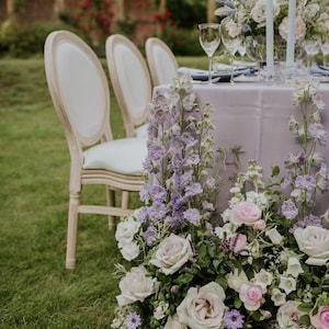 May include: Elegant outdoor dining scene with a lavender tablecloth, white chairs, and floral arrangements. The table is set with glassware and candles. The flowers include roses and delphiniums, creating a romantic ambiance.