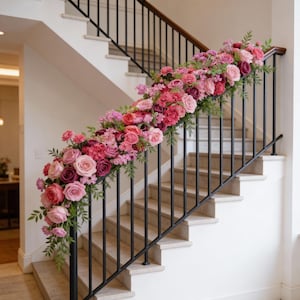 May include: A staircase with white steps and a black metal railing is adorned with a garland of pink and red roses, hydrangeas, and greenery. The floral arrangement cascades down the railing, creating a decorative and elegant display.
