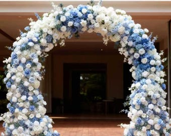 Puede incluir: Un arco floral decorativo compuesto por flores blancas y azules. Las flores están densamente dispuestas, formando una forma llena y redondeada. El arco se encuentra frente a un edificio con suelo de madera.