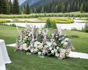 Könnte beinhalten: Ein üppiges Blumenarrangement mit weißen, rosa und lavendelfarbenen Blumen, auf einem grünen Rasen. Die Anordnung befindet sich vor einer malerischen Kulisse aus See und Bergen, mit einem weißen Stoffläufer.