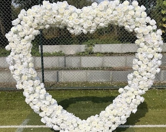 May include: A white heart-shaped floral arch made of artificial white roses. The arch is standing on a green grassy surface.