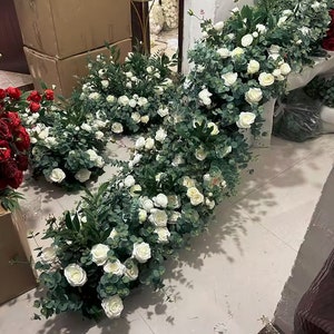 Puede incluir: Una colección de arreglos florales artificiales con rosas blancas y follaje verde. Los arreglos se muestran en un suelo de baldosas de color gris claro.