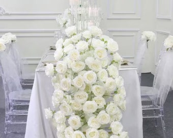 May include: A table setting with a white tablecloth, adorned with cascading white roses. Clear acrylic chairs are visible, along with a candelabra centerpiece. The overall theme suggests a wedding or formal event.