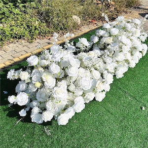 May include: A lush arrangement of white roses and smaller white flowers, possibly baby's breath, creating a dense, floral display. The flowers are arranged on a bed of green artificial turf, suggesting a decorative or event setting.